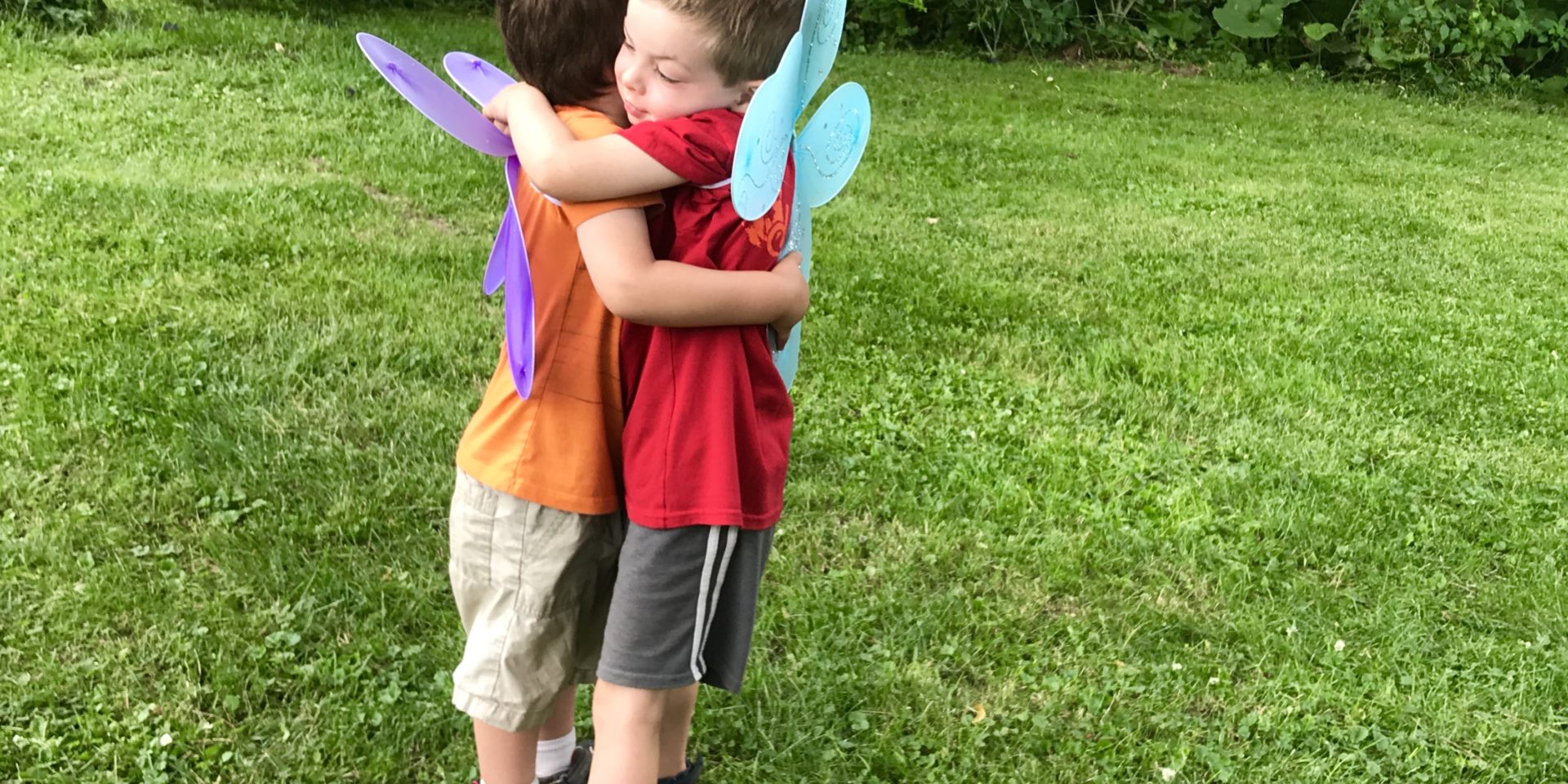 Two children wearing butterfly wings give one another a hug.