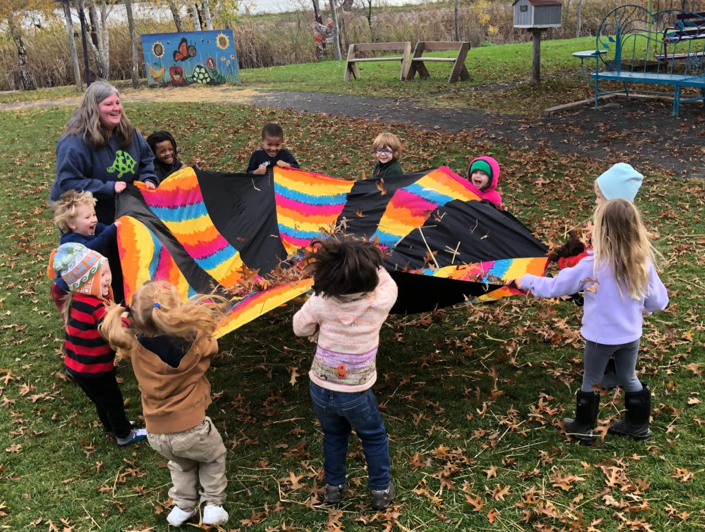 A white teacher and several small children of different races lift a colorful parachute blanket with fallen leaves in the center together. All of them are smiling.