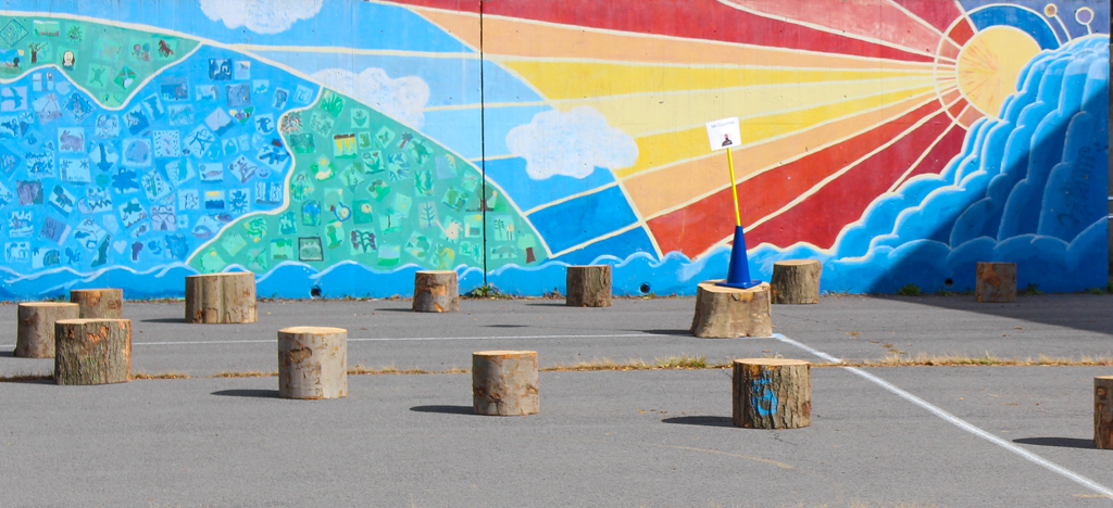 Vibrantly colored mural of a sun and its rays shining on the Earth. Tree stumps placed on the lot in front of it. 
