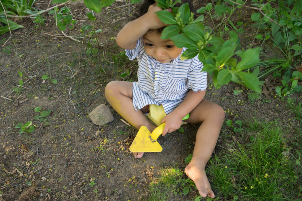 Small child hiding in a bush and sitting in the dirt with a yellow shovel in his hand. 