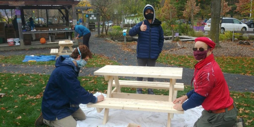 Three folks sanding a natural wood picnic table, one is giving a thumbs up.