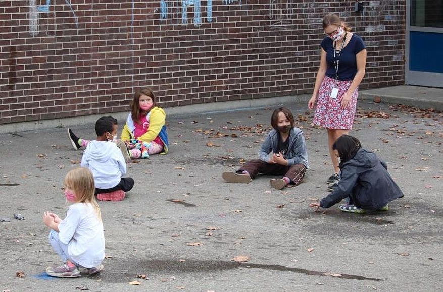 Several seated children on pavement, a teacher stands among them. 