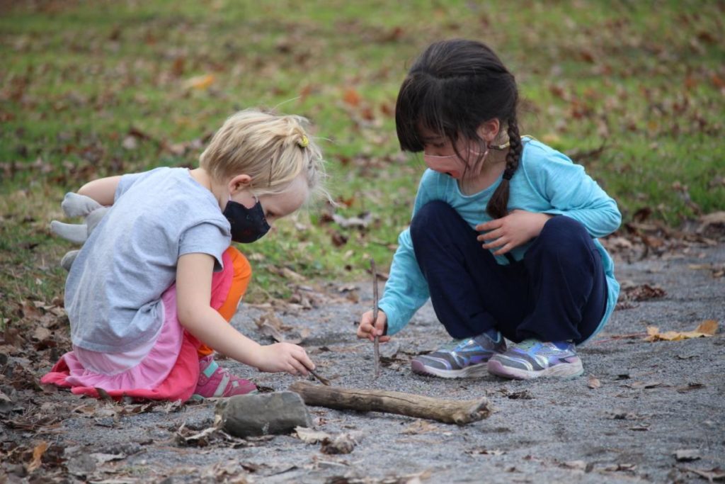 Two younger children, wearing face mask coverings, playing in the dirt with twigs.