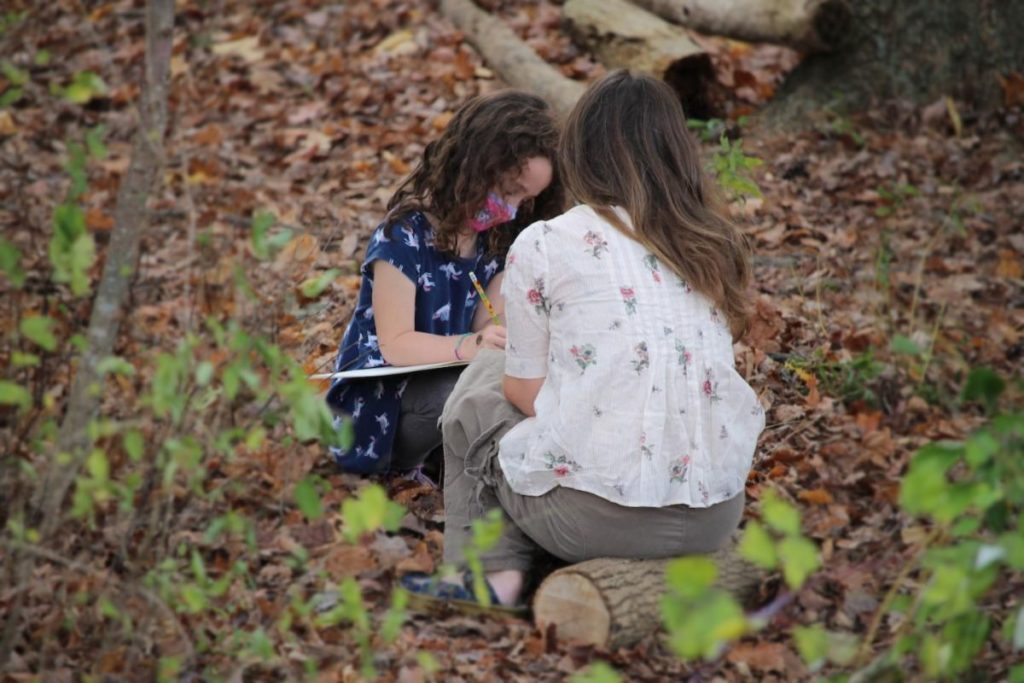 A young student sitting in the leaves intently writing, a teacher sitting on a cut off log seat looks on. 