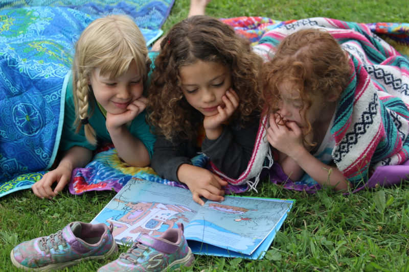 49352235366_744dc9db58_b Three children lay on the grass and read a book together.