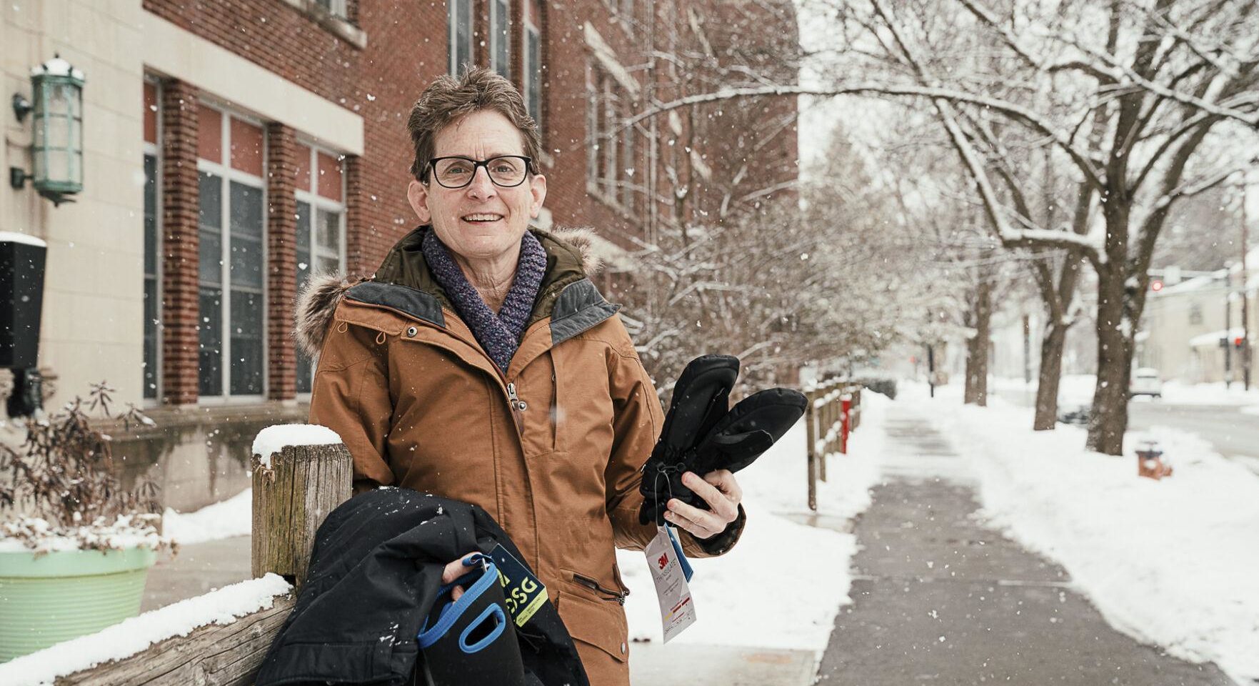 Pale skinned person holds winter gear in their hand while standing outside in snow.