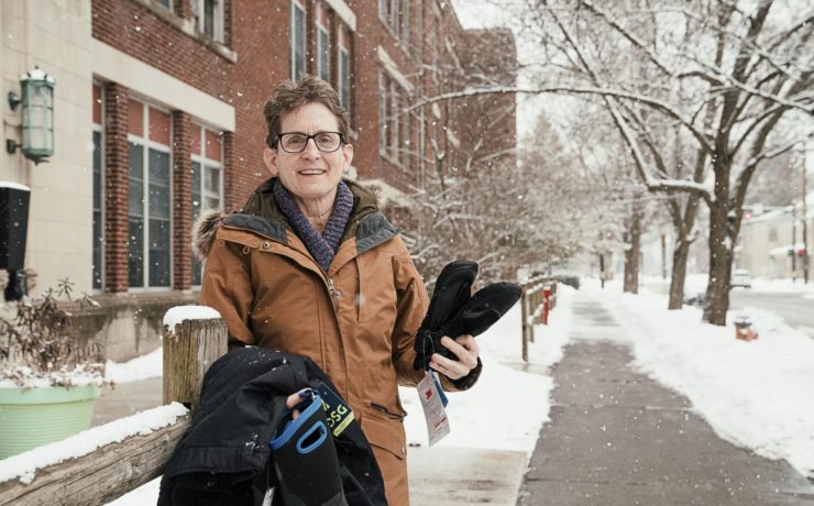 Pale skinned person holds winter gear in their hand while standing outside in snow.