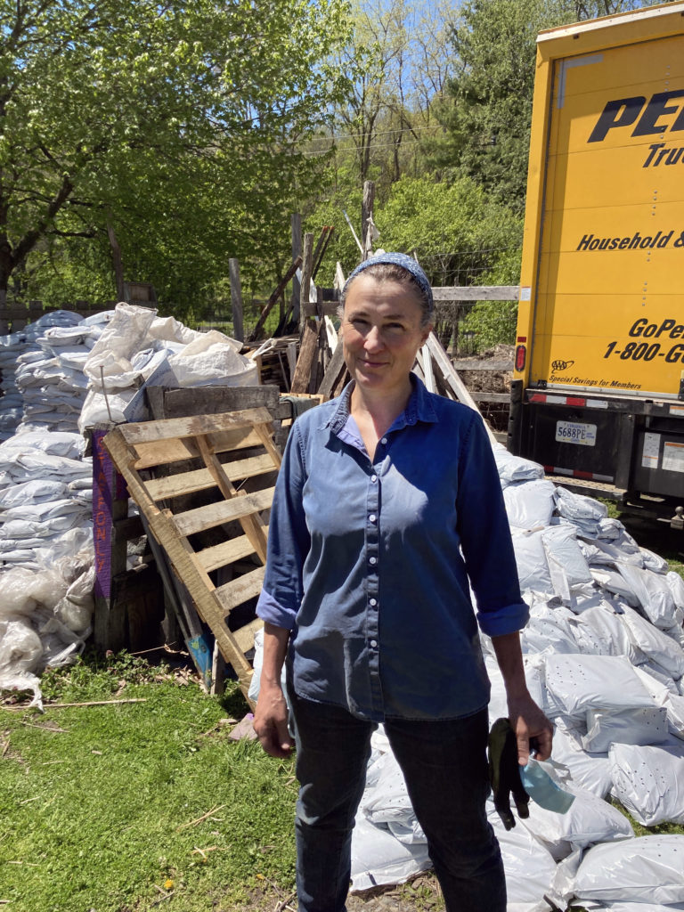 Peggy Arcadi stands in front of a large yellow truck, piles of large white bags full of soil behind them. They are smiling at the camera. 