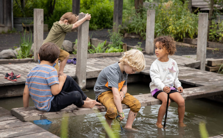 Three preschoolers sit beside one another on the dock of the rice paddy pond.