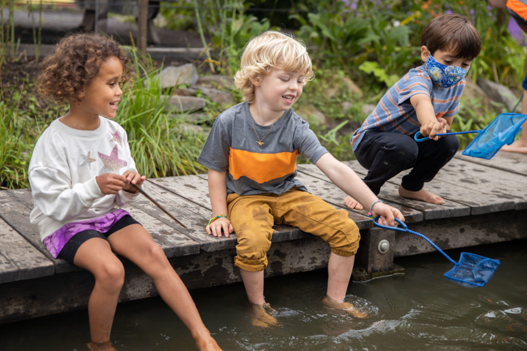 Children playing outside at a shallow pond, seated on a deck, laughing and enjoying themselves. 