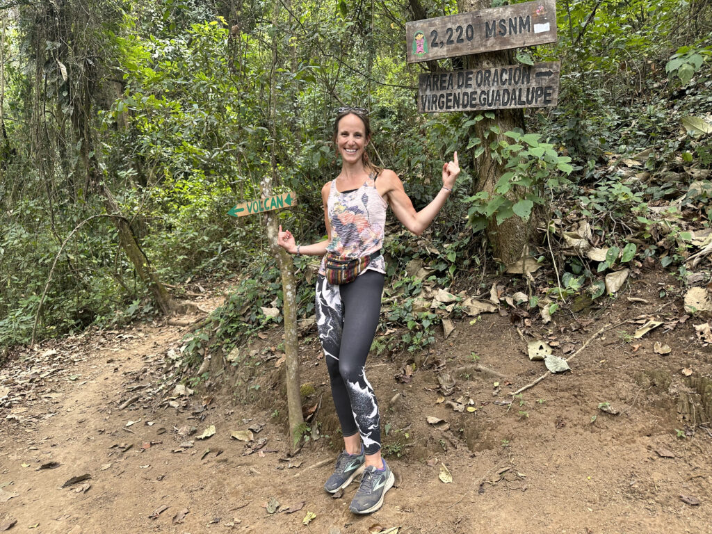 Erin Marteal is outside between two trail wayfinder posts, laughing as she points separately at both. One sign reads "Volcan" and pointing to the left. The other trail sign says, "2,200 MSNM. Area de oracion virgendeguadalupe", while pointing right. 
