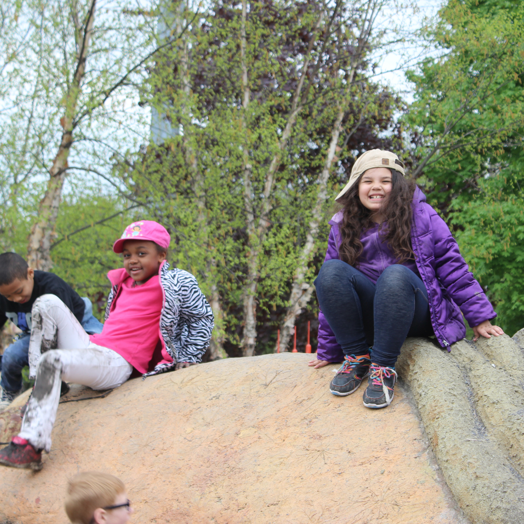 Two kids sit on a giant sculpture of a turtle named Gaia, located in Ithaca Children's Garden. The kids beam at the camera, excited to be high up.