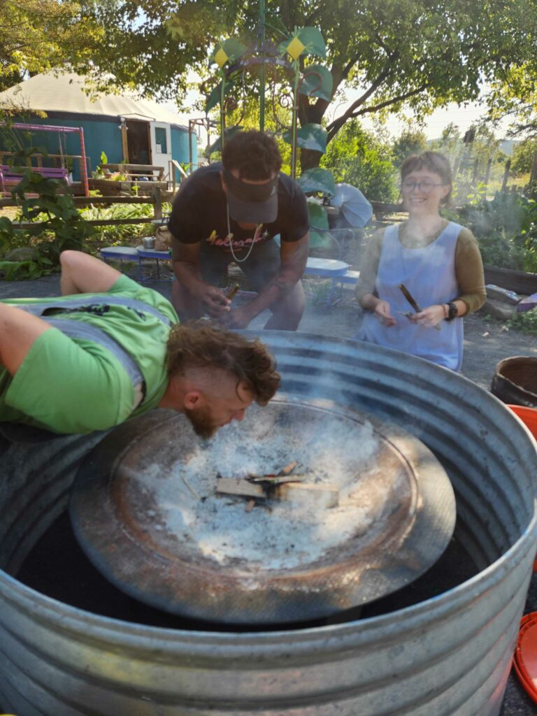 Jason McClevish demonstrates fire safety to two ICG educators at Ithaca Children's Garden. He blows on smoking wood, feeding oxygen to the flames.