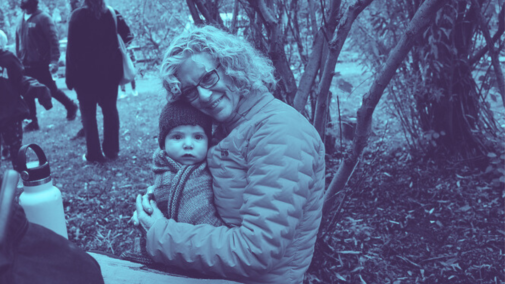 Woman holding a baby smiles at the camera. They are seated on a bench outdoors at Ithaca Children's Garden.