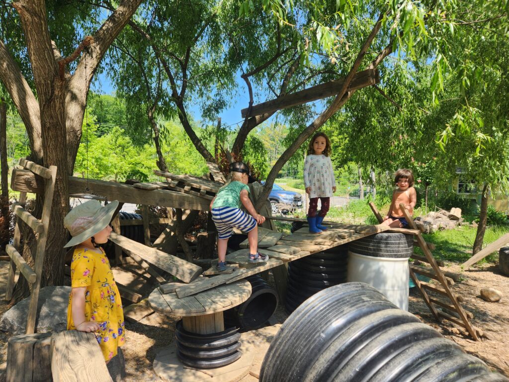 Four kids climb atop a structure made from wood, repurposed wire spools, and ladders, in Ithaca Children's Garden's Hands-On Nature Anarchy Zone.