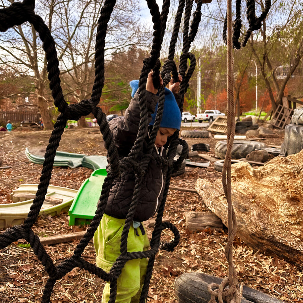 Keith, a Playful Nature Explorer, frowns in frustration as he tries to climb the rope web in ICG's outdoor Hands-On Nature Anarchy Zone.