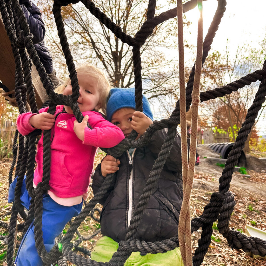 Keith and another child, both Playful Nature Explorers at Ithaca Children’s Garden, are smiling and laughing together on a rope web in the Hands-On Nature Anarchy Zone, surrounded by fall leaves, embodying the joy of childhood adventures.