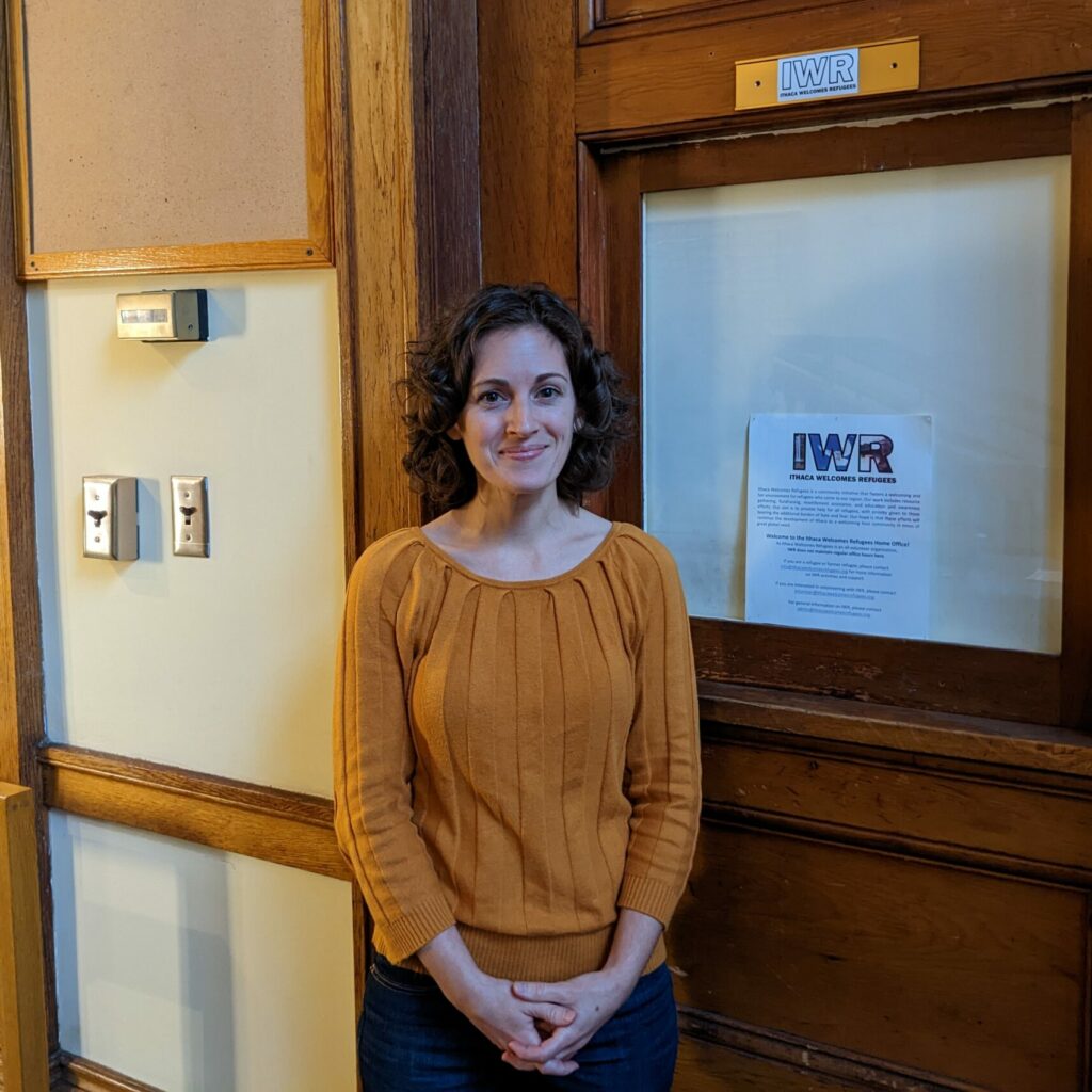 A portrait of Casey Verderosa standing confidently in front of a door labeled 'IWR,' representing the Executive Director of Ithaca Welcomes Refugees. 