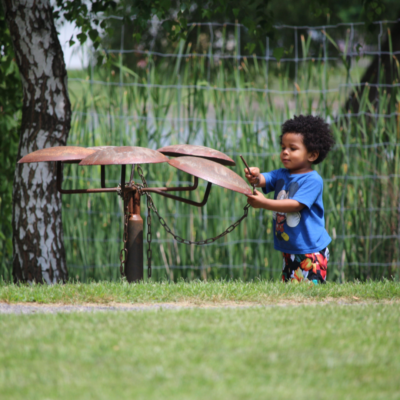 a child stands holding a metal stick that they use to tap on metal cymbals in the outdoors