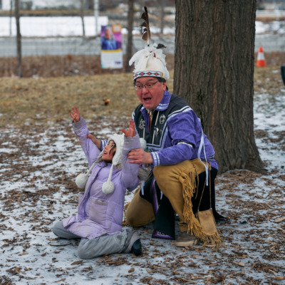 A man in traditional Haudenosaunee dress interacts with a child in winter clothing by raising their arms