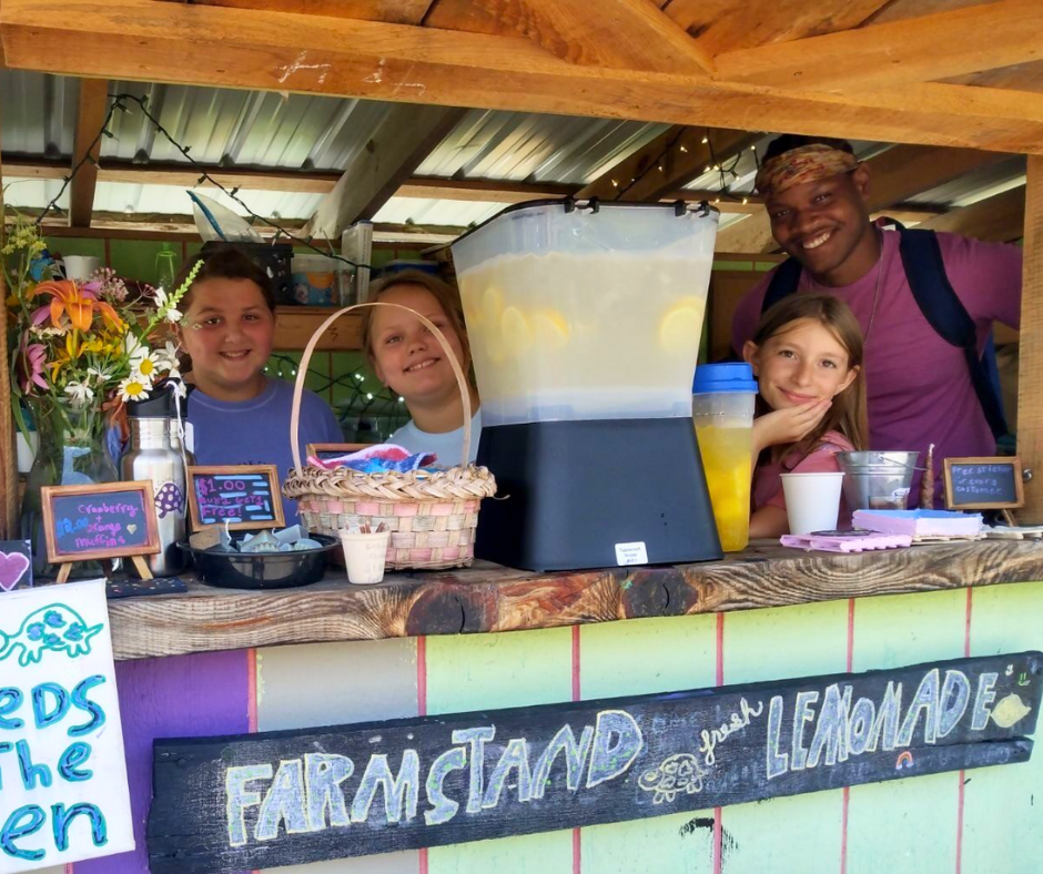 Three children and an educator stand inside a farm stand near lemonade.