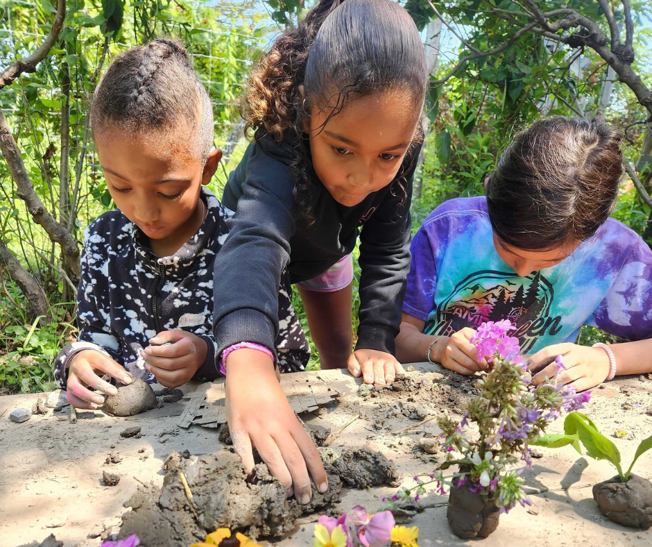 Three children work with dirt and flowers in a garden setting.