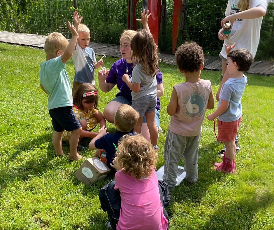 A group of young children stands in the grass around an instructor. Some raise their hands.